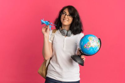 young-pleased-pretty-caucasian-schoolgirl-with-headphones-neck-wearing-glasses-back-bag-holds-plane-globe-looking-up-pink-with-copy-space_141793-62632.jpg