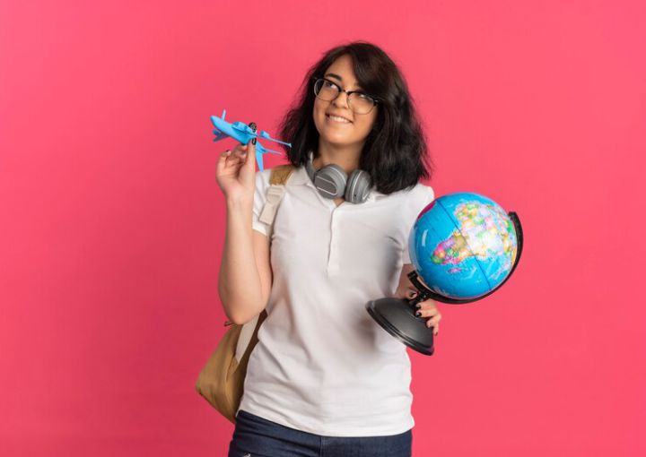 young-pleased-pretty-caucasian-schoolgirl-with-headphones-neck-wearing-glasses-back-bag-holds-plane-globe-looking-up-pink-with-copy-space_141793-62632.jpg